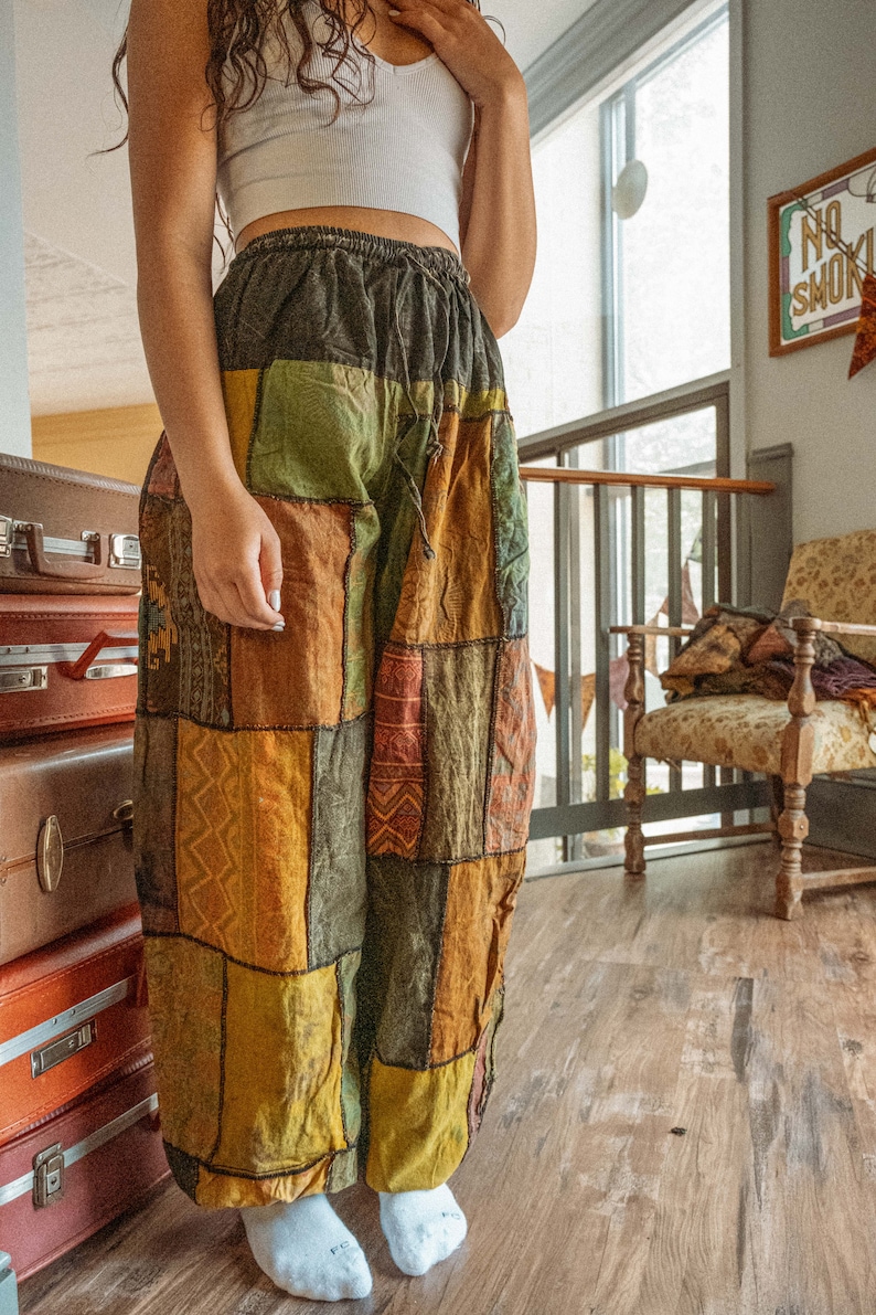May include: A person wearing a white crop top and patchwork pants with a drawstring waist. The pants are made of different fabrics and colors, including brown, green, yellow, and orange. The person is standing in a room with wooden floors and a chair in the background.