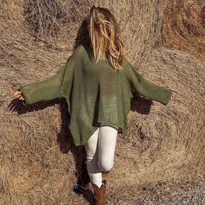 May include: An olive green, oversized knit sweater with long sleeves. The sweater is paired with light-colored pants and brown cowboy boots. The person is standing in front of a large hay bale.