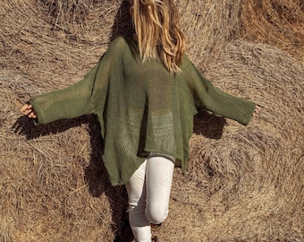 May include: An olive green, oversized knit sweater with long sleeves. The sweater is paired with light-colored pants and brown cowboy boots. The person is standing in front of a large hay bale.