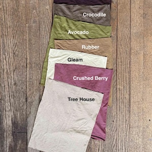 May include: A collection of fabric swatches in various colors, including Black Bean, Crocodile, Avocado, Rubber, Gleam, Crushed Berry, and Tree House. Each swatch has a label with its corresponding color name. The swatches are arranged on a wooden surface.