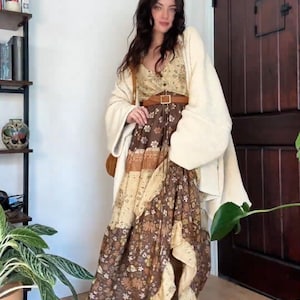 Puede incluir: Una mujer lleva un vestido maxi largo de flores marrones y crema con un cinturón de cuero marrón. Lleva un cárdigan blanco y botas marrones. El vestido tiene una falda de capas y un corpiño ajustado.