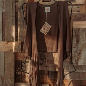 May include: A brown sleeveless top with a tie front detail. The top is hanging on a black hanger against a wooden background. The top has a small tag attached to it that says "Sunshine Shop".