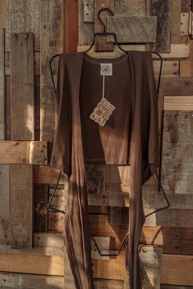 May include: A brown sleeveless top with a tie front detail. The top is hanging on a black hanger against a wooden background. The top has a small tag attached to it that says "Sunshine Shop".