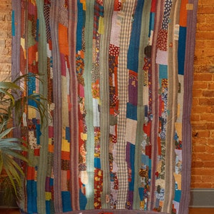 Op de afbeelding: Een kleurrijk patchwork-deken met verticale strepen in tinten blauw, groen, rood en paars. Het deken is gemaakt van verschillende stoffen en patronen, waaronder bloemenprints, ruitjes en stippen.