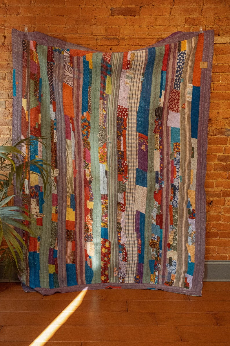 Op de afbeelding: Een kleurrijk patchwork-deken met verticale strepen in tinten blauw, groen, rood en paars. Het deken is gemaakt van verschillende stoffen en patronen, waaronder bloemenprints, ruitjes en stippen.
