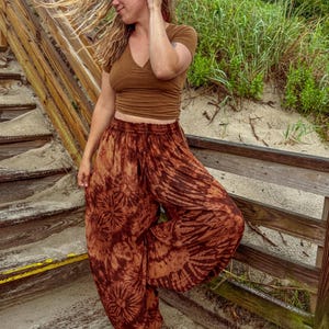 May include: Brown tie-dye harem trousers with a matching brown V-neck crop top. The trousers feature a unique circular and striped pattern. The outfit is worn on a wooden boardwalk near the beach.