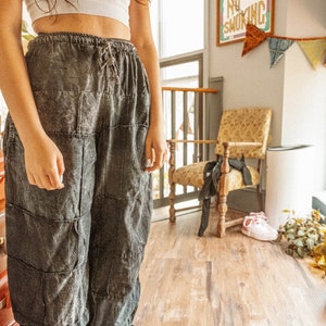 May include: A person wearing a white crop top and dark gray patchwork pants. The pants have a drawstring waist and are loose fitting. The person is standing in a room with a wooden floor and a chair in the background. There is a sign that says "No Smoking" on the wall.