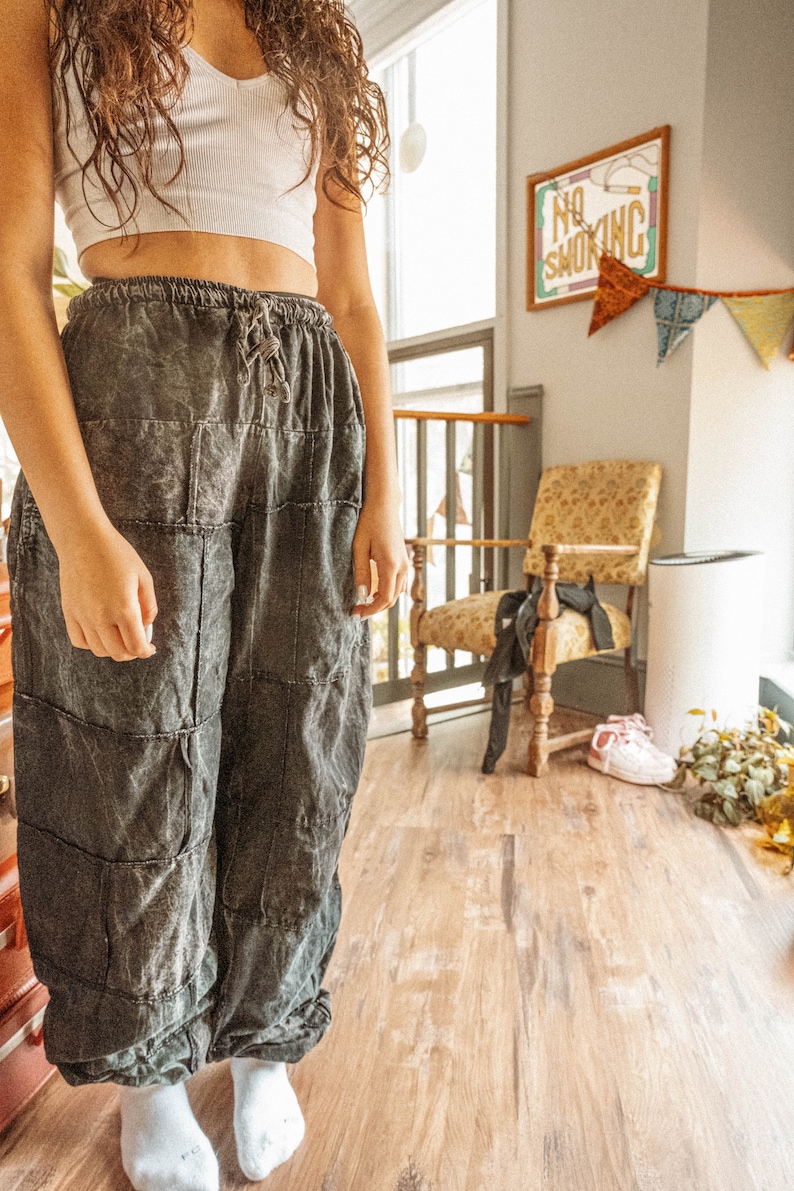 May include: A person wearing a white crop top and dark gray patchwork pants. The pants have a drawstring waist and are loose fitting. The person is standing in a room with a wooden floor and a chair in the background. There is a sign that says "No Smoking" on the wall.