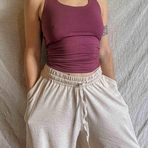 K&ouml;nnte beinhalten: Ein dunkelpflaumenfarbenes Neckholder-Top mit rundem Ausschnitt und gerafftem Stoff an der Taille, kombiniert mit einer locker sitzenden, hellbeigen melierten Hose mit elastischem Bund und passendem Kordelzug mit dunklen Metallenden.