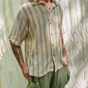 Puede incluir: Una camisa de manga corta a rayas verticales beige y verde oliva, con botones y un bolsillo en el pecho. Se combina con pantalones verde oliva con bolsillos grandes. El conjunto es informal y relajado.
