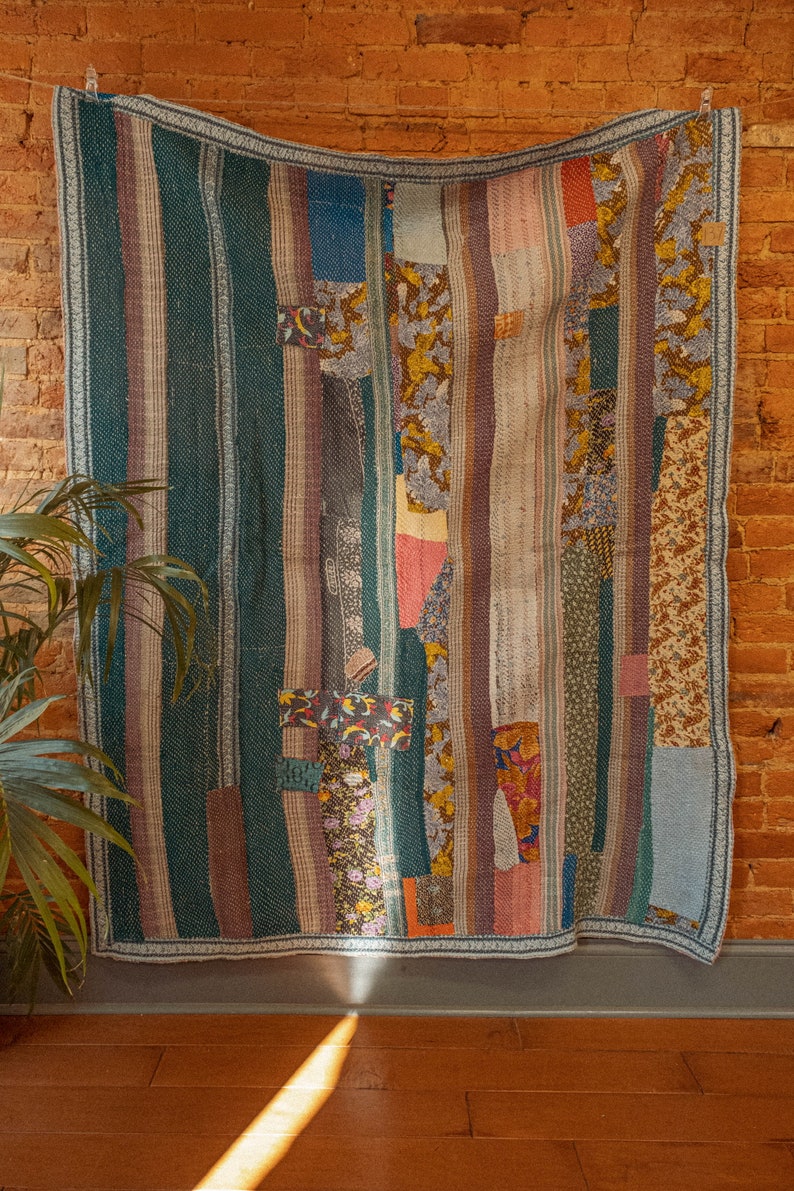 Op de afbeelding: Een patchworkdeken met een verscheidenheid aan kleuren en patronen, waaronder groen, blauw, roze en geel. De deken is gemaakt van verschillende stoffen die in een gestreept patroon aan elkaar zijn genaaid.