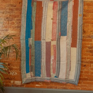 May include: A colorful patchwork quilt with stripes of blue, red, white, and green fabric. The quilt is hanging on a brick wall.
