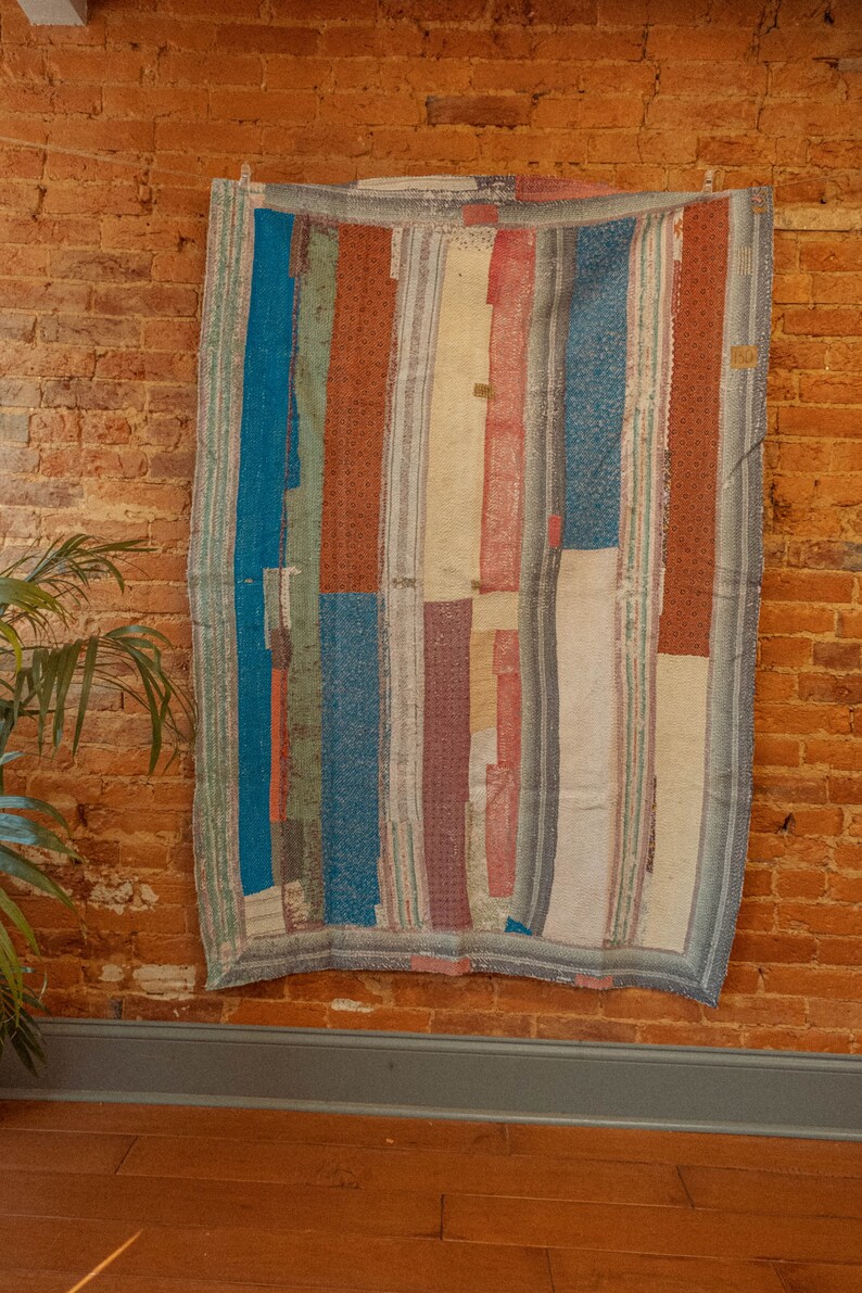 May include: A colorful patchwork quilt with stripes of blue, red, white, and green fabric. The quilt is hanging on a brick wall.