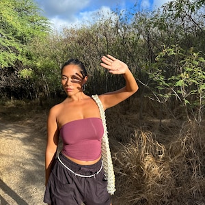 May include: A woman wearing a burgundy tube top and black shorts with a white drawstring. She is carrying a cream-colored bag with a beaded strap. The background features a sunny outdoor scene with trees and a blue sky.