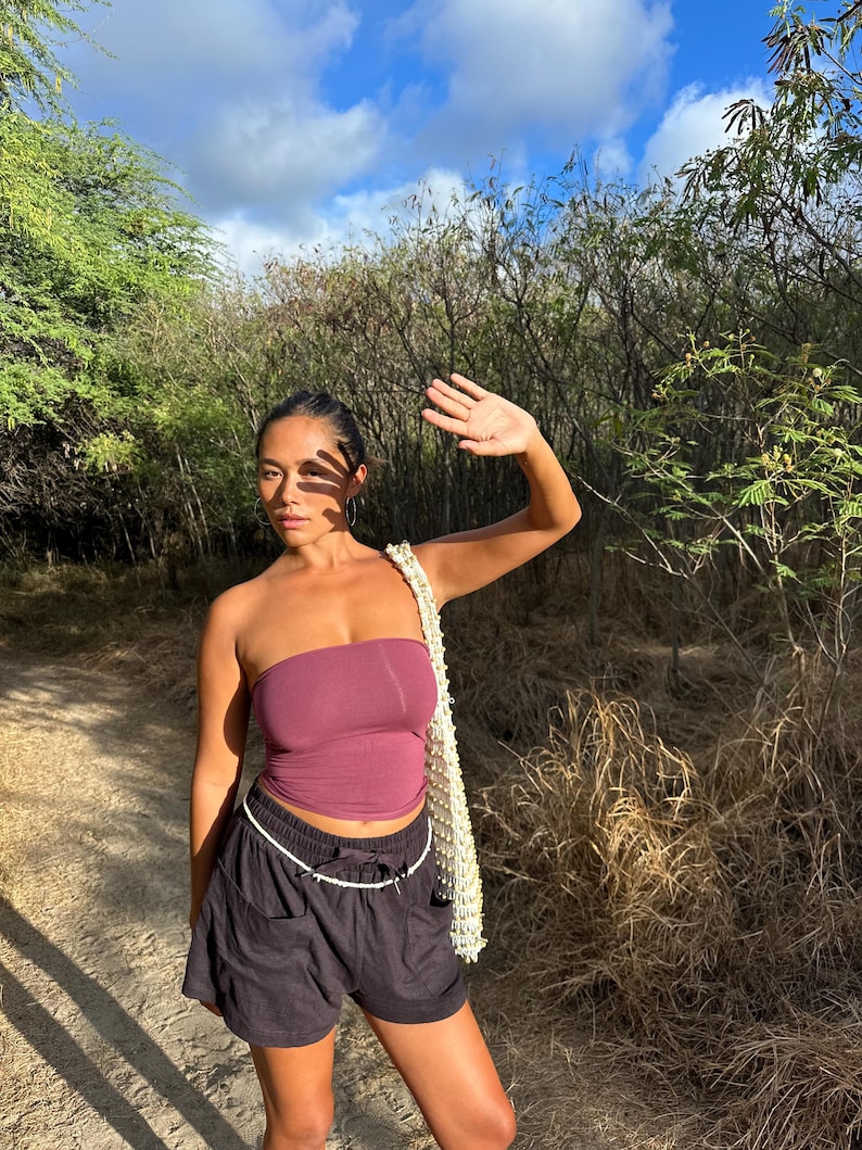 May include: A woman wearing a burgundy tube top and black shorts with a white drawstring. She is carrying a cream-colored bag with a beaded strap. The background features a sunny outdoor scene with trees and a blue sky.