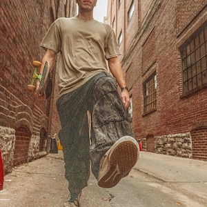 May include: A person walks down a brick alleyway carrying a skateboard. They are wearing a tan t-shirt, dark pants, and white sneakers.