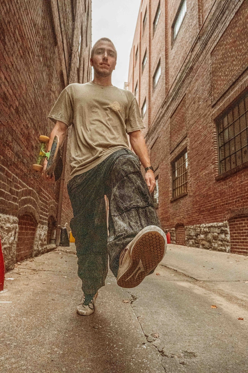 May include: A person walks down a brick alleyway carrying a skateboard. They are wearing a tan t-shirt, dark pants, and white sneakers.