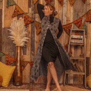 May include: A woman wearing a long, dark gray vest with a floral pattern over a black turtleneck dress. She is standing in front of a wooden wall with a string of colorful pennants hanging from it.