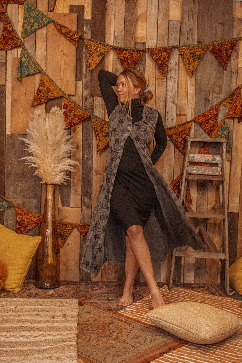 May include: A woman wearing a long, dark gray vest with a floral pattern over a black turtleneck dress. She is standing in front of a wooden wall with a string of colorful pennants hanging from it.