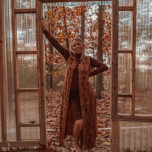 May include: A woman wearing a long brown dress with a floral pattern stands in a doorway of a wooden structure with a clear plastic roof. The background is a forest with fall foliage.