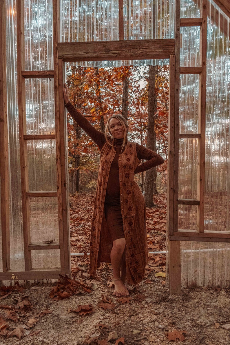 May include: A woman wearing a long brown dress with a floral pattern stands in a doorway of a wooden structure with a clear plastic roof. The background is a forest with fall foliage.