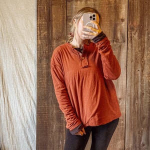 May include: Rust-colored long-sleeve Henley shirt with a three-button placket. The shirt has dark gray cuffs and a dark gray collar. The person in the image is wearing black pants.