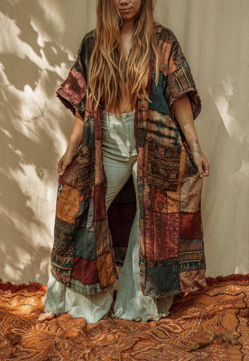 以下が含まれることがあります： 茶色、赤、青、金など、さまざまな色と模様が施された長いパッチワークの着物。着物は前で開いており、その下に白いズボンが見えます。