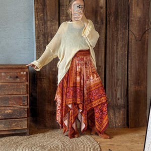 May include: A woman wearing a cream-colored, loose-fitting sweater and a long, flowing skirt with a vibrant floral pattern in shades of red, orange, and pink. The skirt has an asymmetrical hemline. The outfit is paired with brown boots.