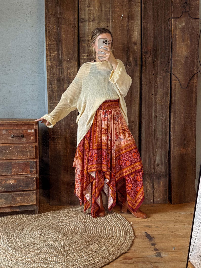 May include: A woman wearing a cream-colored, loose-fitting sweater and a long, flowing skirt with a vibrant floral pattern in shades of red, orange, and pink. The skirt has an asymmetrical hemline. The outfit is paired with brown boots.