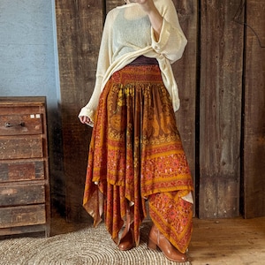May include: A long, flowing skirt with a vibrant orange and brown paisley pattern. The skirt has a tiered design and a gathered waistband. The outfit is paired with a cream-colored sweater and brown ankle boots. The skirt's length is approximately 36 inches.