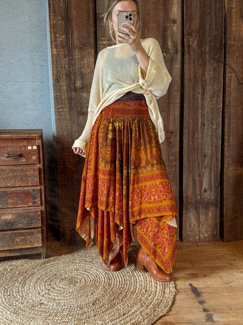 May include: A long, flowing skirt with a vibrant orange and brown paisley pattern. The skirt has a tiered design and a gathered waistband. The outfit is paired with a cream-colored sweater and brown ankle boots. The skirt's length is approximately 36 inches.