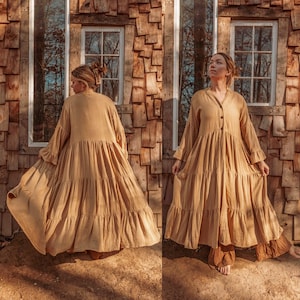 以下が含まれることがあります： 長い袖とボタン留めのフロントを持つ、長い、ティアードのライトブラウンのドレスを着た女性。ドレスはゆったりとしたフィット感で、柔らかく流れるような生地で作られています。女性は窓のある木製の壁の前に立っています。
