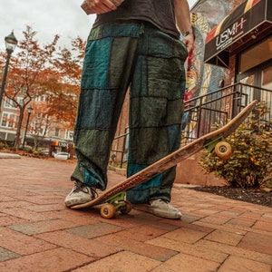 May include: A person wearing a black shirt and patchwork pants is standing on a skateboard with a brown deck and green wheels. The person is wearing a brown and white striped bag slung over their shoulder. The skateboard is on a brick walkway.