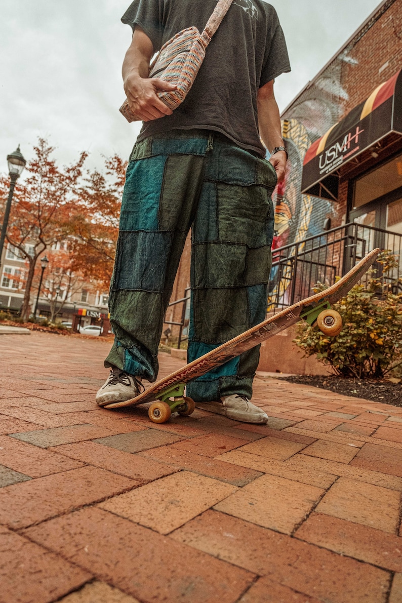 May include: A person wearing a black shirt and patchwork pants is standing on a skateboard with a brown deck and green wheels. The person is wearing a brown and white striped bag slung over their shoulder. The skateboard is on a brick walkway.