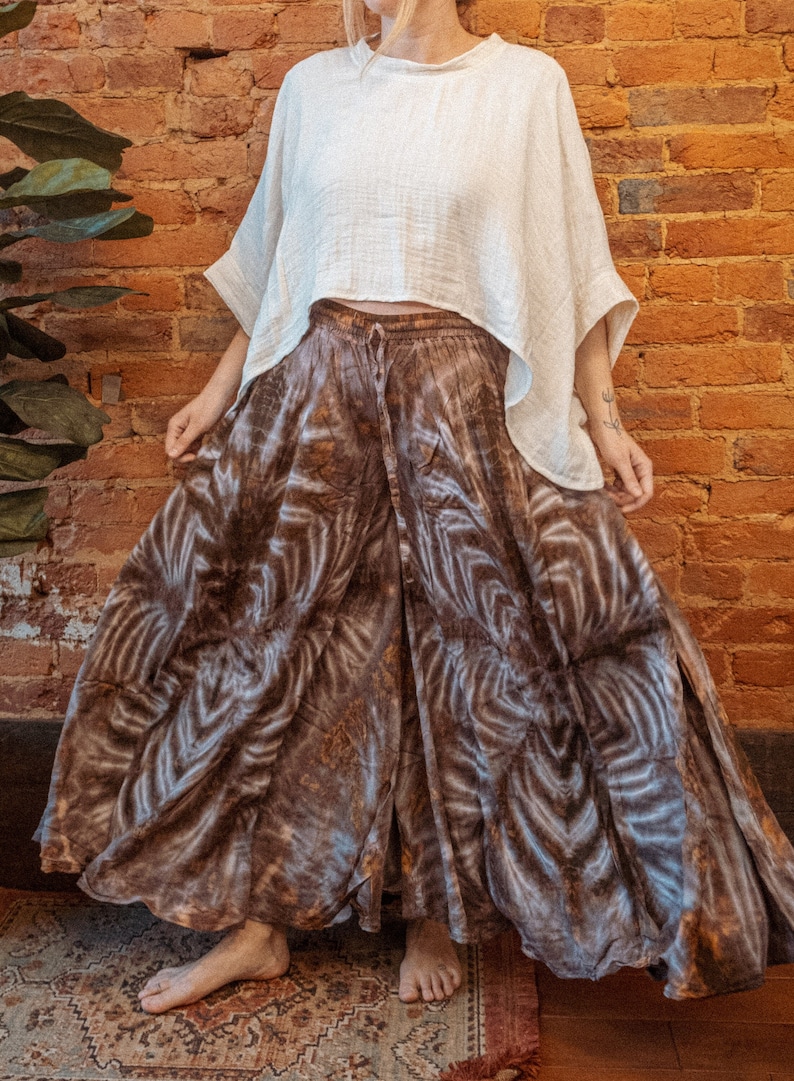 May include: A woman wearing a white, loose-fitting top and a long, brown and white tie-dye skirt. The skirt has a flowing, wide-leg silhouette. The woman is standing in front of a brick wall.