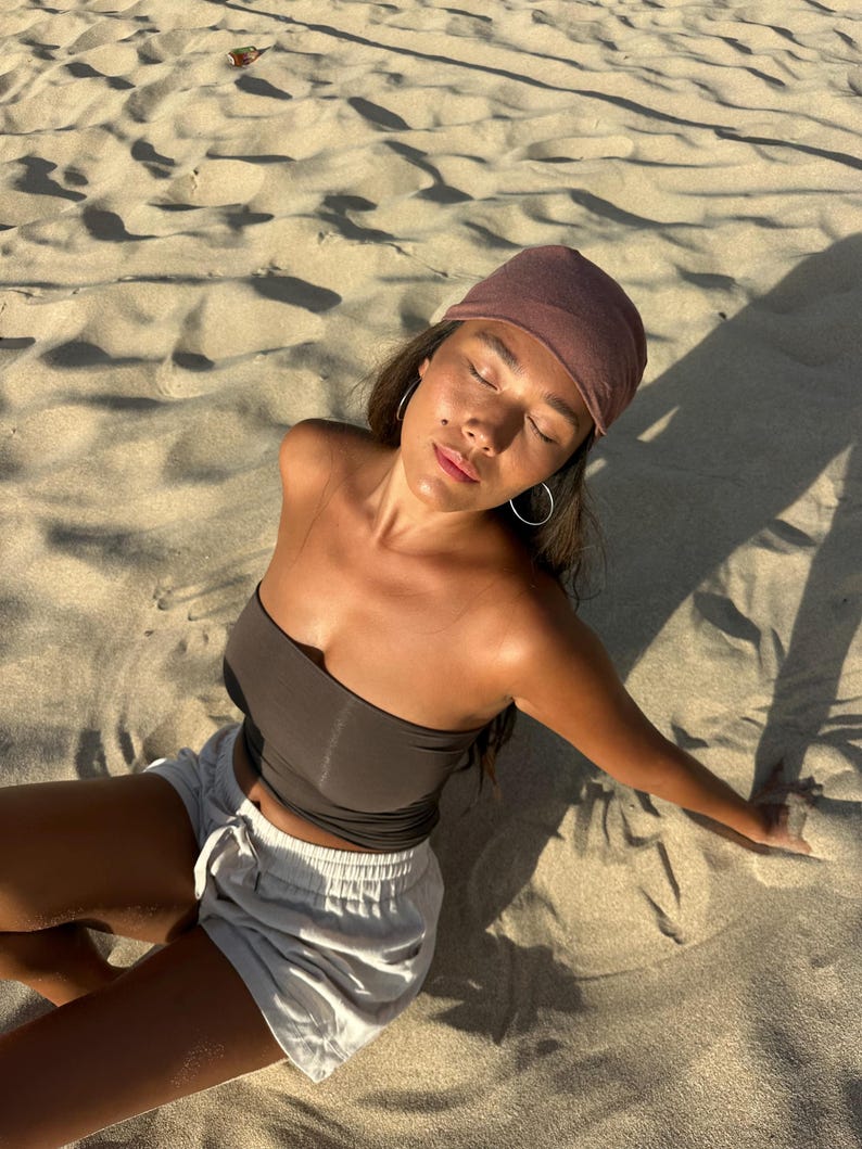 May include: A person wearing a brown tube top, light gray shorts, and a maroon headscarf, sitting on a sandy beach. The person is wearing silver hoop earrings. The image is taken in natural sunlight.