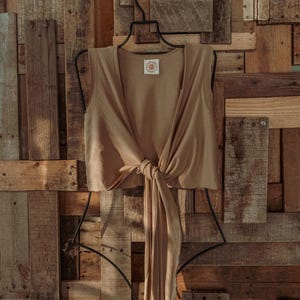 May include: A brown sleeveless wrap top with a tie at the waist, hanging on a black hanger against a rustic wooden background.