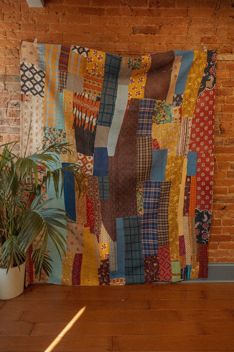 May include: A colorful patchwork quilt with a variety of fabrics and patterns in shades of blue, yellow, red, and brown. The quilt is hanging on a brick wall.