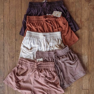 May include: An assortment of casual shorts in various colors, including deep purple, rust, cream, and muted pink. Each pair features an elastic waistband with a drawstring and side pockets. The shorts are arranged on a wooden surface.
