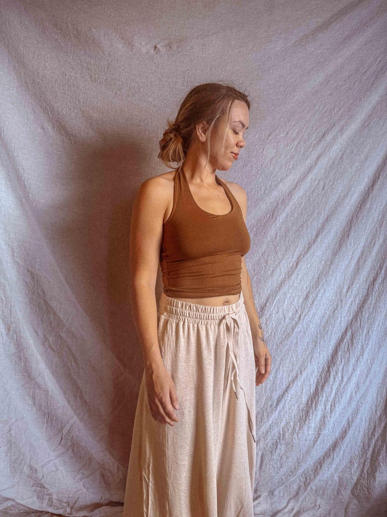 K&ouml;nnte beinhalten: Ein einfarbig braunes, &auml;rmelloses Neckholder-Top mit rundem Ausschnitt, kombiniert mit einem hellbeigen Rock mit hoher Taille, elastischem Bund und einem funktionalen Kordelzug auf der Vorderseite, das ein schlichtes zweiteiliges Outfit bildet.