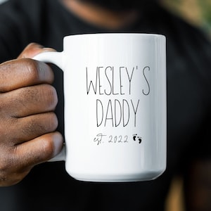 May include: White ceramic mug with black text that reads "Wesley's Daddy est. 2022" with two small footprints below the text.