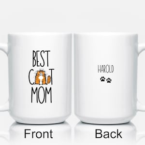 May include: White ceramic coffee mug with a black design on the front that says "Best Cat Mom" with a cartoon cat image. The back of the mug has the name "Harold" and two paw prints.