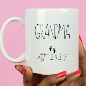 May include: White ceramic mug with the word "GRANDMA" in black script, accompanied by two small black footprints and the text "est. 2023". The mug has a curved handle and is held against a pink background.