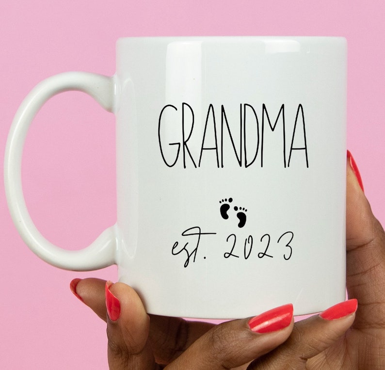 May include: White ceramic mug with the word "GRANDMA" in black script, accompanied by two small black footprints and the text "est. 2023". The mug has a curved handle and is held against a pink background.