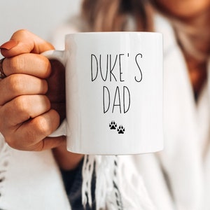 May include: White ceramic mug with black text that reads "Duke's Dad" and two black paw prints.