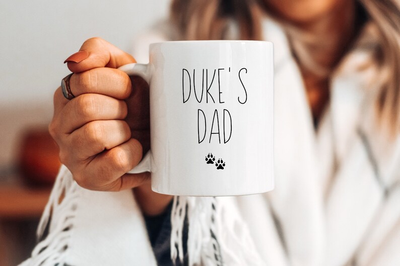 May include: White ceramic mug with black text that reads "Duke's Dad" and two black paw prints.