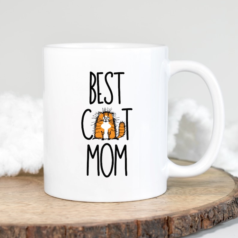 May include: White ceramic coffee mug with a black and white graphic of a cat and the text "BEST CAT MOM".
