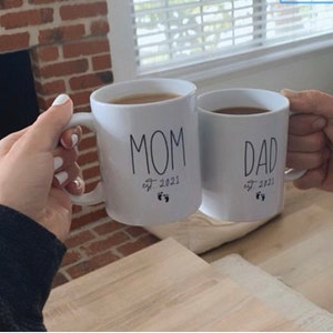 May include: Two white ceramic mugs with black lettering. The mug on the left says "MOM est. 2021" with a footprint symbol. The mug on the right says "DAD est. 2021" with a footprint symbol.