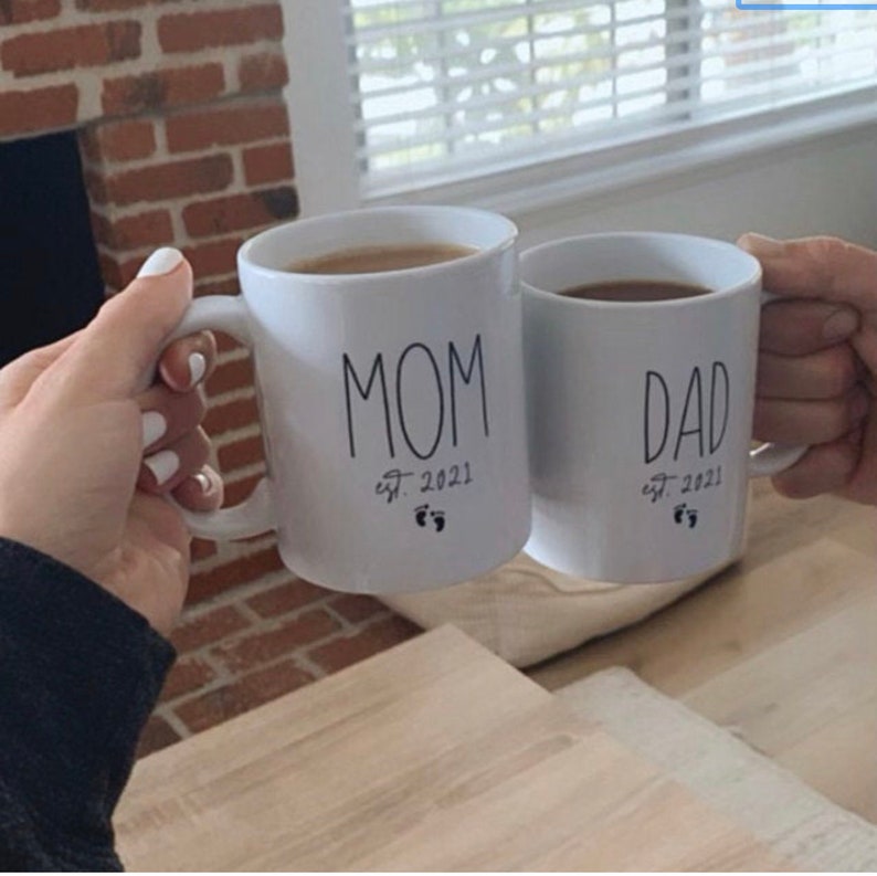 May include: Two white ceramic mugs with black lettering. The mug on the left says "MOM est. 2021" with a footprint symbol. The mug on the right says "DAD est. 2021" with a footprint symbol.