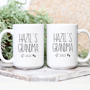 Op de afbeelding: Twee witte keramische mokken met zwarte tekst die "Hazel's Grandma est. 2022" met een paar voetafdrukken zegt.